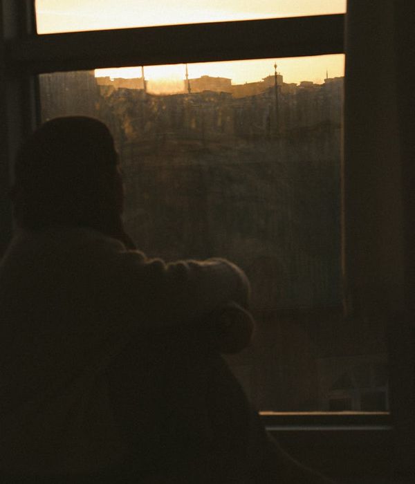 Woman looking thoughtfully out a large window at a vibrant city skyline during sunset.